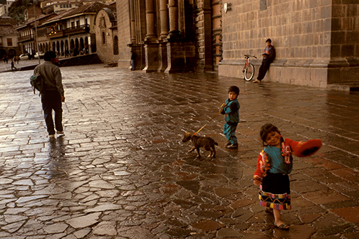 A young girl in traditional Indian dress dances through Cuzco's Plaza de las Armas as her brother hangs onto the family dog at rear. The kids were put on display for their mother to attract alms from passers-by. © Kevin Moloney, 1996.